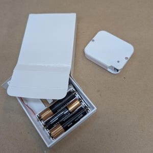 Könnte beinhalten: Eine weiße rechteckige Box mit einem Klappdeckel, geöffnet, um vier AAA-Batterien zu zeigen. Ein separater weißer rechteckiger Deckel ist ebenfalls vorhanden. Die Batterien sind von der Marke Duracell.