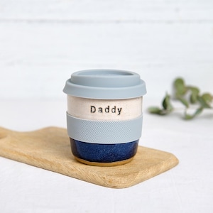 May include: A ceramic travel mug with a blue silicone lid and a matching textured band. The mug is white with the word "Daddy" printed on it, and has a dark blue base. It sits on a wooden cutting board.