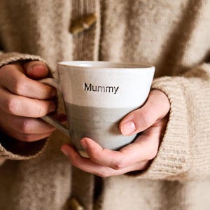 May include: A ceramic mug with the word "Mummy" printed on the upper white section. The lower section is a muted grey. The mug is held by two hands, suggesting a cosy, warm beverage experience.