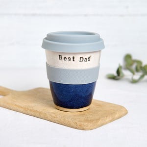 May include: A ceramic travel mug with a light blue lid and a matching textured band. The mug is white with the words "Best Dad" printed on it, and the bottom half is a deep blue. The mug sits on a wooden cutting board.