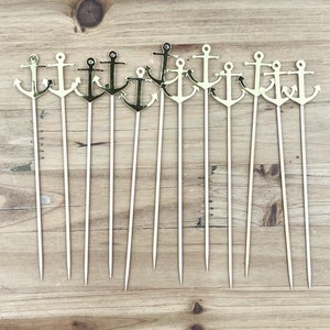 May include: A set of 12 gold anchor cocktail picks. The picks are made of wood and have a gold-colored anchor at the top. The anchors are all facing the same direction.