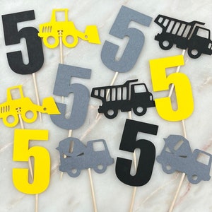 May include: Set of six construction-themed cake toppers. Each topper features a number five in yellow, black, or gray, with a different construction vehicle in the same color scheme. The vehicles include a dump truck, a cement mixer, and a bulldozer.
