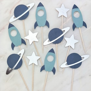 May include: A set of ten blue and silver space-themed cupcake toppers. The toppers feature rockets, planets with rings, and stars.
