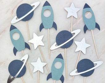 May include: A set of ten blue and silver space-themed cupcake toppers. The toppers feature rockets, planets with rings, and stars.