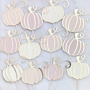 May include: A collection of pastel-colored pumpkin-shaped decorations on white sticks. The pumpkins have a layered design with gold trim and swirling vines. The color palette includes shades of pink, white, and gold, perfect for fall-themed events.