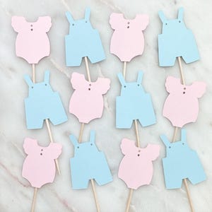 May include: A collection of baby shower cupcake toppers. The toppers are in the shape of baby onesies and overalls. The onesies are pink, and the overalls are light blue. Each topper is attached to a wooden stick.