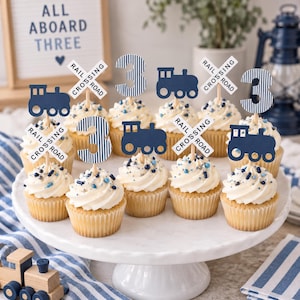 May include: A collection of cupcakes on a white pedestal, each topped with a decorative pick featuring a dark blue train, a white railroad crossing sign, or a blue and white striped number three. The cupcakes have white frosting and blue sprinkles.