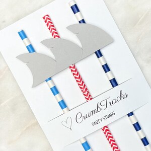 May include: A set of three paper straws with shark fin toppers. The straws are red and white striped, blue and white striped, and red and white chevron. The package has a heart and the text "Crumb Tracks Party Straws".