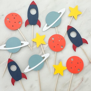 May include: A set of 10 space-themed cupcake toppers. The toppers feature rockets, planets, stars, and moons in shades of blue, red, yellow, and white. The toppers are perfect for a space-themed party.