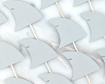 May include: Light gray paper shark fin cupcake toppers on wooden sticks. The fins are arranged in a pattern against a white marble background. These toppers are ideal for a shark-themed party or event.