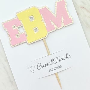 May include: A cake topper with the letters "EBM" in pink and yellow, outlined with gold glitter. The topper is attached to a wooden skewer and includes the text "CrumbTracks CAKE TOPPER".