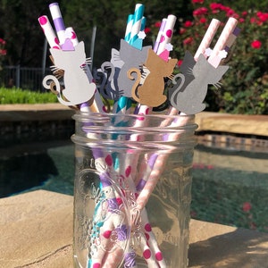 May include: A glass jar filled with colorful striped paper straws with cat-shaped paper cutouts attached to the tops. The straws are pink, purple, teal, and white with pink polka dots. The cat cutouts are gray, brown, and white.