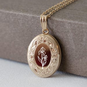 May include: A gold-toned oval locket pendant suspended from a matching delicate chain. The locket features an intricate embossed floral border surrounding a central smooth panel engraved with a white line drawing of an iris flower.