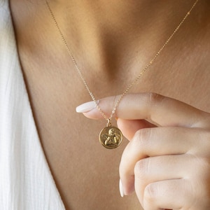Może przedstawiać: Złoty naszyjnik z łańcuszkiem i małym, okrągłym wisiorem z wytłoczonym wzorem anioła.