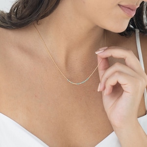 Peut inclure: Un collier délicat en chaîne dorée avec de petites perles turquoise. Le collier est porté par une personne avec un haut blanc.