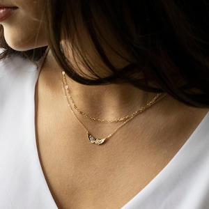 Pu&ograve; includere: Una collana a catena d'oro con un delicato ciondolo a forma di ala. La collana &egrave; sovrapposta a una seconda catena pi&ugrave; sottile.