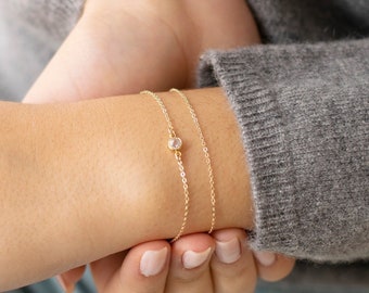 May include: A delicate gold chain bracelet with a small clear gemstone. The bracelet is layered on a person's wrist.