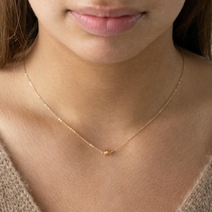 Könnte beinhalten: Eine zarte goldene Halskette mit einem kleinen, runden Anhänger. Die Halskette wird über einem neutralfarbenen Pullover getragen. Die Kette ist fein und der Anhänger ist zentriert, was einen minimalistischen und eleganten Look erzeugt. Der Schmuck ist schlicht und edel.