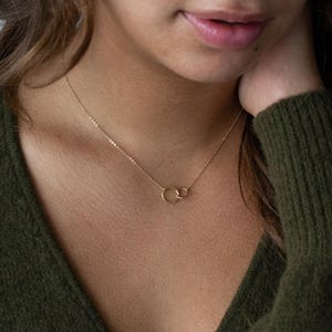 Peut inclure: Un collier délicat en or composé de deux petits pendentifs circulaires entrelacés avec une finition martelée. La fine chaîne en or poli est reliée aux anneaux qui reposent sur la clavicule. Le métal présente un ton chaud et réfléchissant.