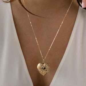 Könnte beinhalten: Eine goldene Halskette mit einem herzförmigen Anhänger. Der Anhänger hat ein filigranes Design und eine kleine Öffnung in der Mitte.