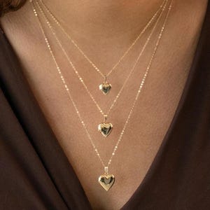 Könnte beinhalten: Eine mehrreihige goldene Halskette mit drei herzförmigen Anhängern. Die Kette wird von einer Person mit braunen Haaren und einem braunen Oberteil getragen.