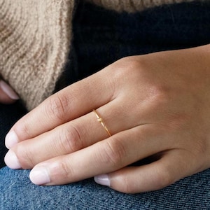 May include: A fine gold ring with a simple, understated design. The ring is slender and has a delicate, textured pattern. The ring is worn on a hand, with a neutral-toned cardigan and blue jeans in the background.