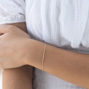 Può includere: Un braccialetto a catena dorata con piccole perline. Il braccialetto è indossato al polso di una persona che indossa una camicetta bianca e jeans blu.