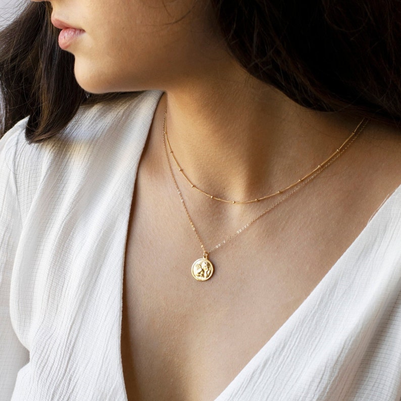 Może przedstawiać: Złoty naszyjnik z delikatnym wisiorem z wypukłym wzorem. Naszyjnik jest nałożony na drugą, cieńszą złotą łańcuszek.