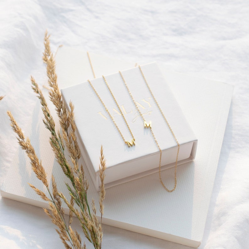 May include: Two gold necklaces with the letter "M" charms in a white box with the word "MIAN" printed on it. The box is sitting on a white surface with a sprig of dried grass.