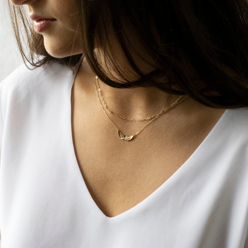 Pu&ograve; includere: Una collana a catena d'oro con un delicato ciondolo a forma di ala. La collana &egrave; sovrapposta a una seconda catena pi&ugrave; sottile.