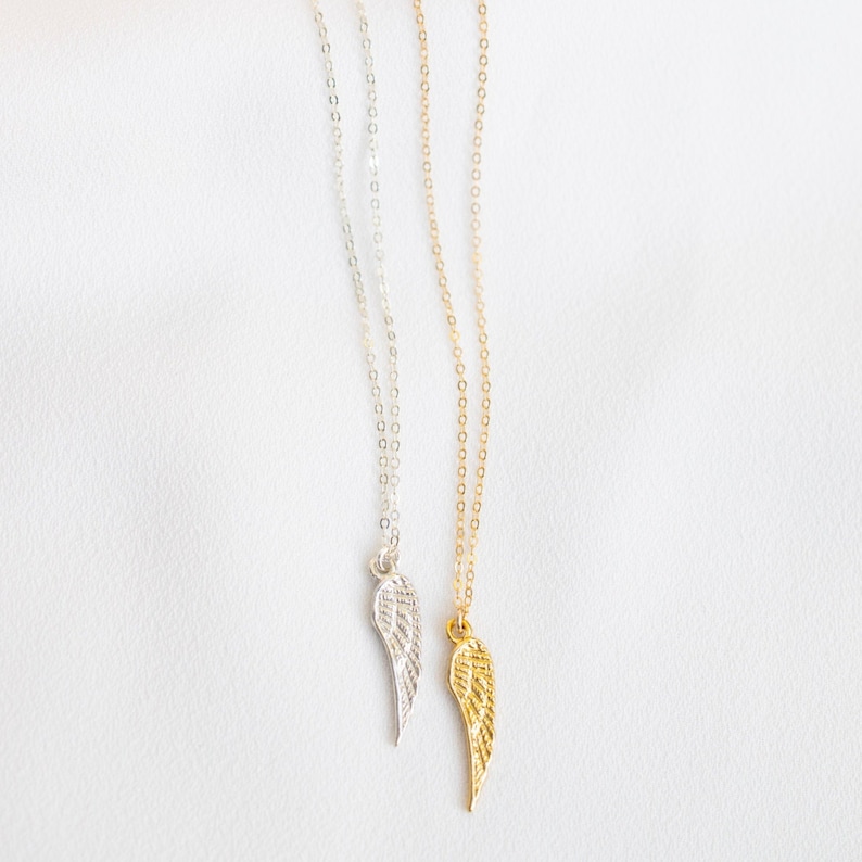 May include: Two delicate necklaces with silver and gold chains and small angel wing pendants. The wings are detailed with a textured design.