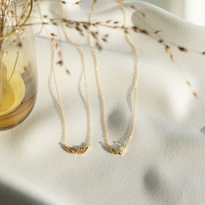 Pu&ograve; includere: Due collane d'oro con catene delicate e piccoli ciondoli a forma di ali d'angelo su uno sfondo di tessuto bianco.