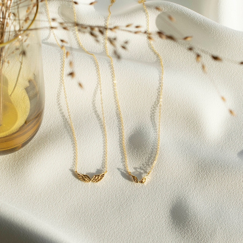 Pu&ograve; includere: Due collane d'oro con catene delicate e piccoli ciondoli a forma di ali d'angelo su uno sfondo di tessuto bianco.