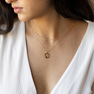 Może przedstawiać: Złoty naszyjnik z delikatnym wisiorem przedstawiającym dwa anioły w reliefie. Naszyjnik jest nałożony na cieńszy złoty łańcuszek.