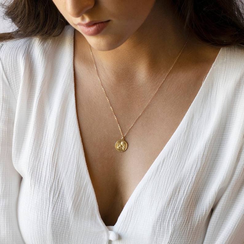 Może przedstawiać: Złoty naszyjnik z łańcuszkiem i małym, okrągłym wisiorem z ozdobnym wzorem. Naszyjnik nosi osoba w białej koszuli.