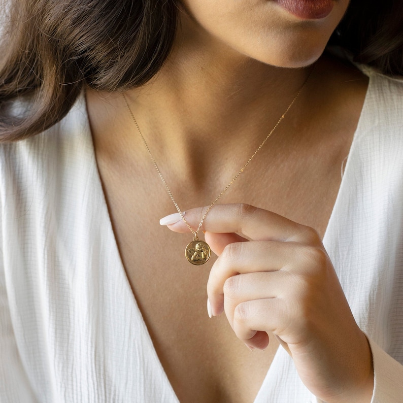 Może przedstawiać: Złoty naszyjnik z łańcuszkiem i małym, okrągłym wisiorem z wytłoczonym wzorem anioła.