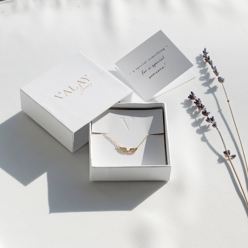 Pu&ograve; includere: Una scatola regalo bianca con le parole "VALAY Jewelry" stampate sulla parte superiore. La scatola &egrave; aperta e contiene una delicata collana d'oro con un ciondolo a forma di ali d'angelo. Un piccolo biglietto bianco con il testo "un regalo speciale - per una persona speciale!" &egrave; visibile sullo sfondo.