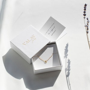 May include: A white jewelry box with the text "VALAY Jewelry" on the lid. The box is open and contains a gold necklace with a small letter "C" pendant. The box is sitting on a white surface with a sprig of lavender in the background.