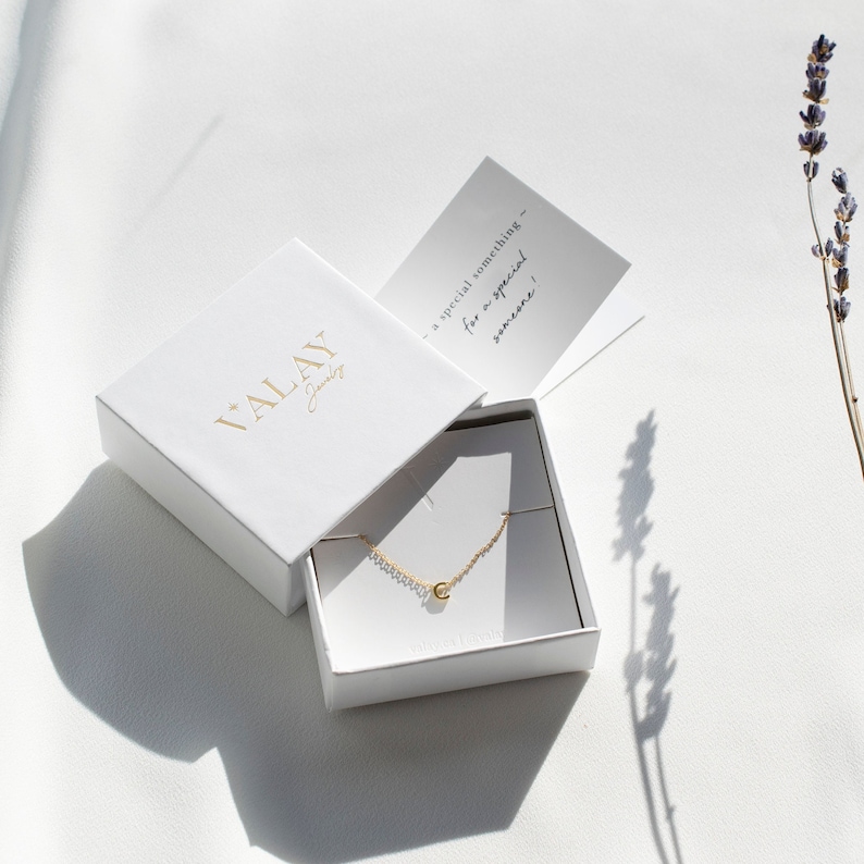 May include: A white jewelry box with the text "VALAY Jewelry" on the lid. The box is open and contains a gold necklace with a small letter "C" pendant. The box is sitting on a white surface with a sprig of lavender in the background.