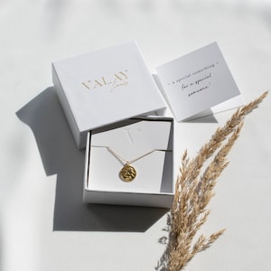Może przedstawiać: Złoty naszyjnik z małym, okrągłym wisiorkiem w białym pudełku. Pudełko jest otwarte, a naszyjnik leży w środku. Na pudełku widnieje napis "Valay Jewelry". Na tle widać kartkę z tekstem "a special something - for a special someone!"