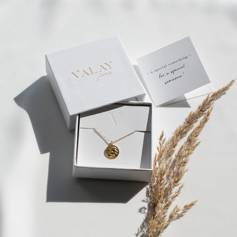 Może przedstawiać: Złoty naszyjnik z małym, okrągłym wisiorkiem w białym pudełku. Pudełko jest otwarte, a naszyjnik leży w środku. Na pudełku widnieje napis "Valay Jewelry". Na tle widać kartkę z tekstem "a special something - for a special someone!"