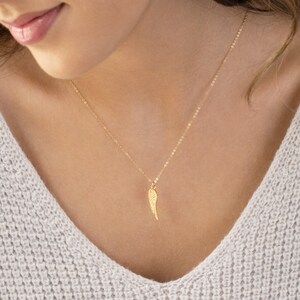 May include: A gold chain necklace with a delicate gold wing pendant. The necklace is worn by a person with a white sweater.
