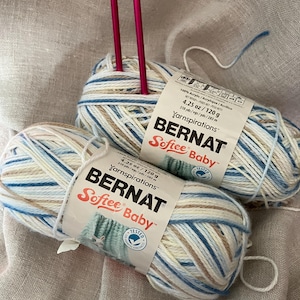 Pode incluir: Dois novelos de fio Bernat Softee Baby numa combinação de cores azul, bege e branco. Cada etiqueta indica 120 g / 4,25 oz. Duas agulhas de tricot cor-de-rosa brilhante estão inseridas no novelo superior.