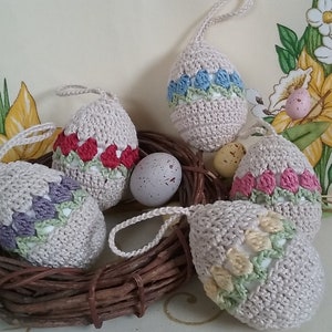 May include: Five crocheted Easter eggs in a bird's nest. The eggs are decorated with different colored flowers. The eggs are beige with purple, red, blue, pink, and yellow flowers.