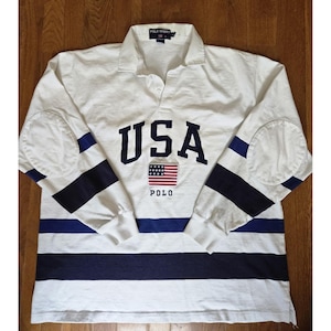May include: White long-sleeve shirt with blue and white stripes and elbow patches. The shirt features the word "USA" in navy blue block letters, an American flag graphic, and the word "POLO".