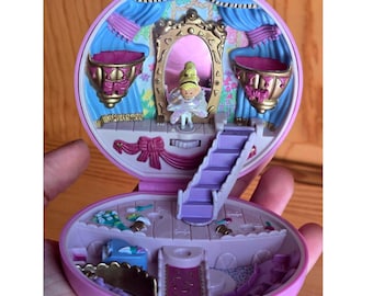 Puede incluir: Un set de juego redondo de plástico rosa que se abre para revelar una escena de salón de baile en miniatura. El interior cuenta con un espejo central con marco dorado, cortinas azules, una pequeña figura con vestido blanco y una escalera.