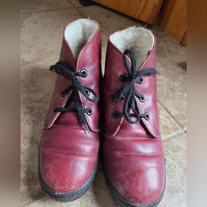 May include: A pair of burgundy leather ankle boots with black laces. The boots have a round toe and a black sole. The interior is lined with a white, fluffy material. The boots are designed for cold weather.
