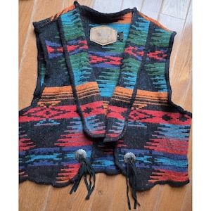 Puede incluir: Un chaleco estampado con un diseño del suroeste. El chaleco presenta patrones geométricos rojos, naranjas, azules y verdes sobre un fondo gris oscuro. Tiene cierres de concho plateados con flecos de cuero negro.