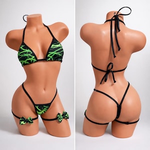 May include: Black bikini set with a green lightning bolt pattern. The set includes a halter top, thong-style bottoms, and garter straps with green bow accents. The bikini is displayed on a mannequin.