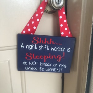 May include: A navy blue wooden door sign with white and red text, and a red and white polka dot ribbon. The sign reads "Shhh.... A night shift worker is Sleeping! do NOT knock or ring unless it's URGENT."